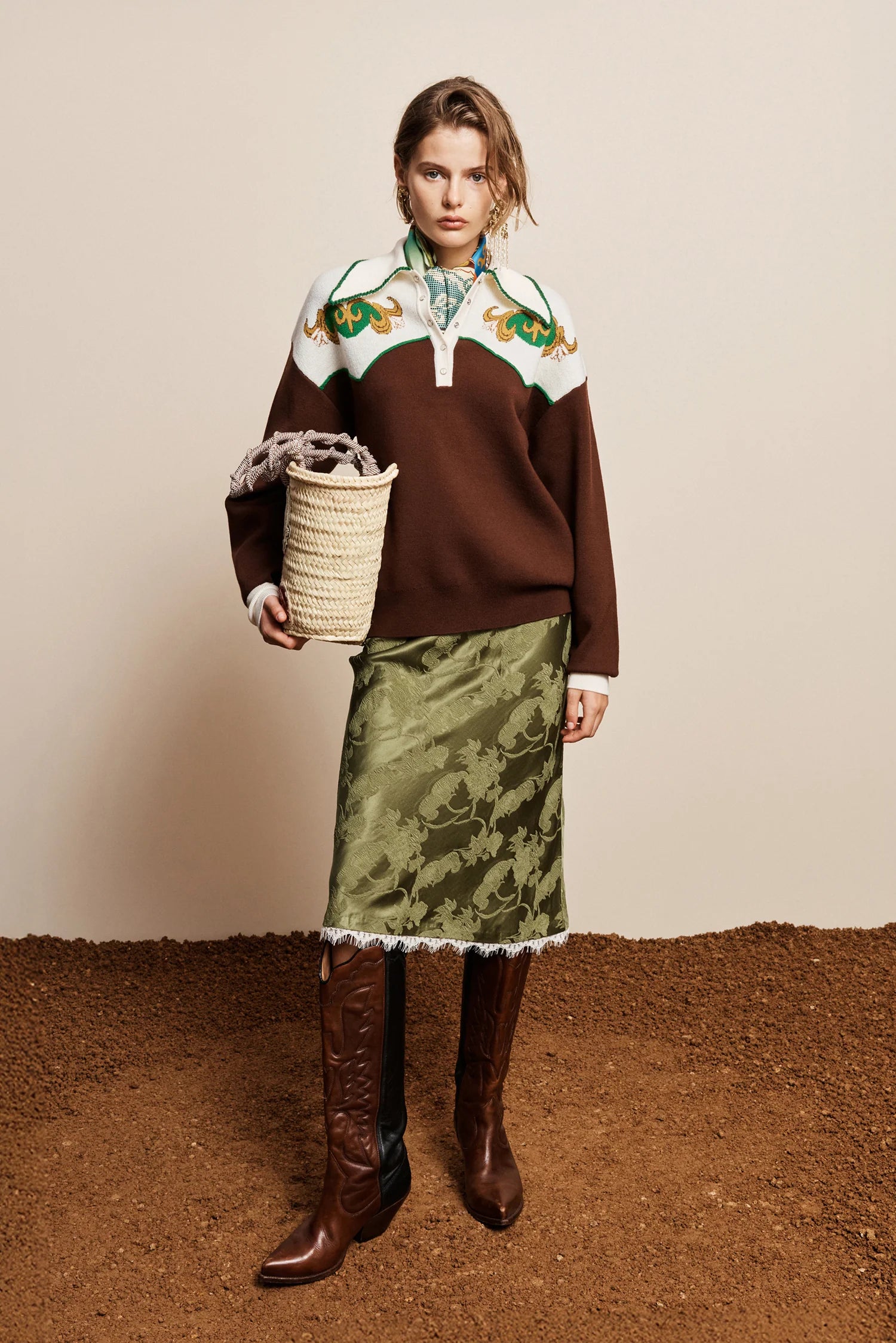 A model stands against a beige wall and brown ground, wearing a brown knit sweater with a white yoke and green floral embroidery, a green patterned skirt with white fringe, and brown knee-high boots. She holds a woven basket.