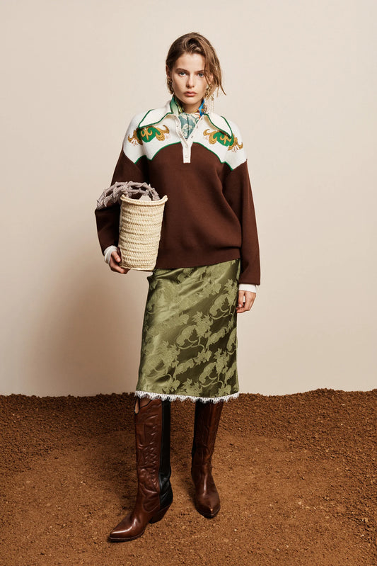 A model stands against a beige wall and brown ground, wearing a brown knit sweater with a white yoke and green floral embroidery, a green patterned skirt with white fringe, and brown knee-high boots. She holds a woven basket.