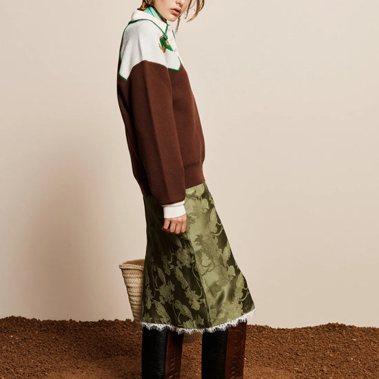 A model is shown from the side wearing a color-blocked brown and white sweater, an olive green patterned midi skirt with white fringe, and brown leather knee-high boots, standing on a textured brown surface against a beige background.