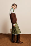 A model is shown from the side wearing a color-blocked brown and white sweater, an olive green patterned midi skirt with white fringe, and brown leather knee-high boots, standing on a textured brown surface against a beige background.