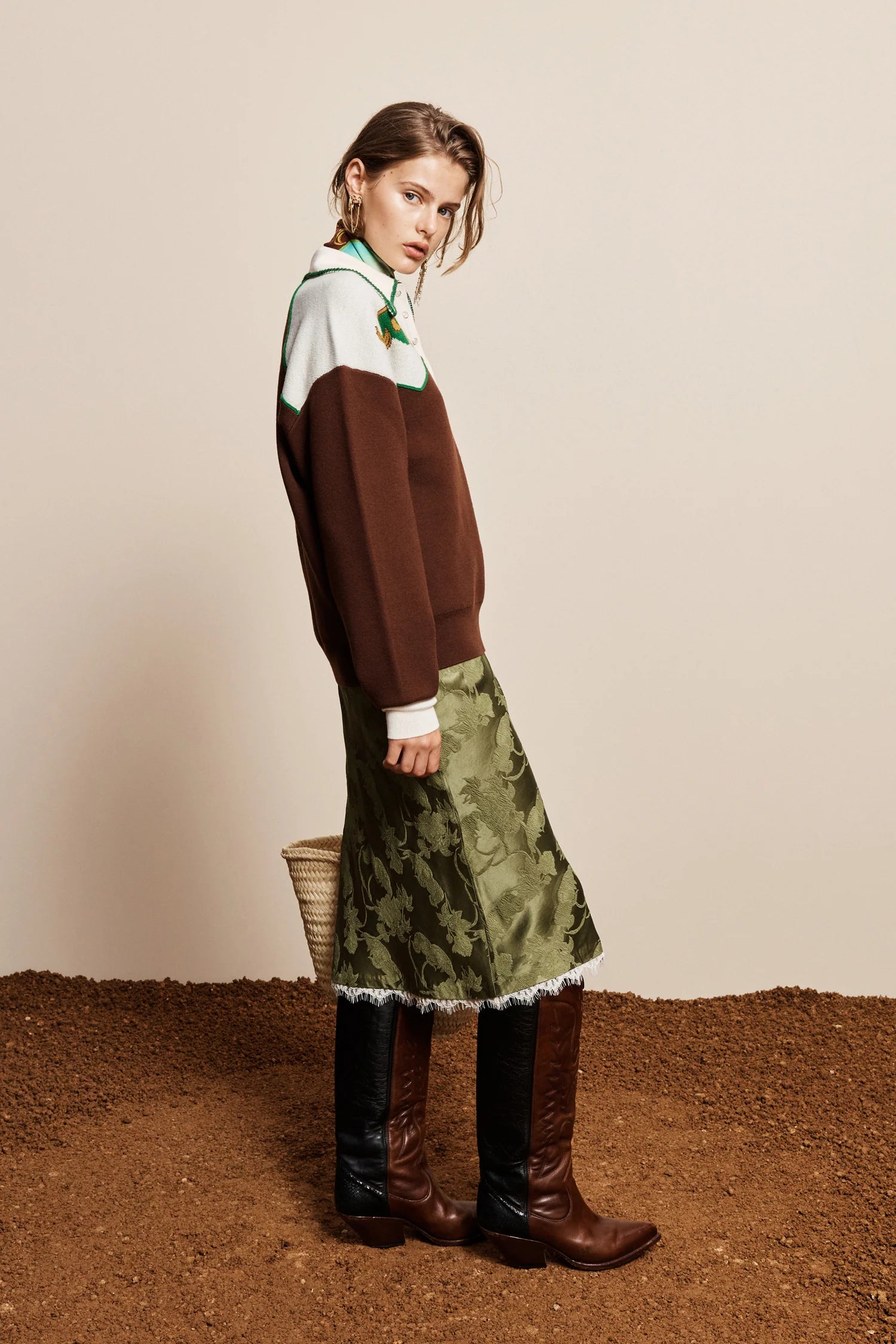 A model is shown from the side wearing a color-blocked brown and white sweater, an olive green patterned midi skirt with white fringe, and brown leather knee-high boots, standing on a textured brown surface against a beige background.