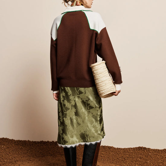 A woman stands with her back to the camera wearing a colorblock knit sweater in brown, white, and green, paired with an olive green patterned skirt, black boots, and holding a woven straw bag against a beige wall and brown floor.