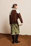 A woman stands with her back to the camera wearing a colorblock knit sweater in brown, white, and green, paired with an olive green patterned skirt, black boots, and holding a woven straw bag against a beige wall and brown floor.