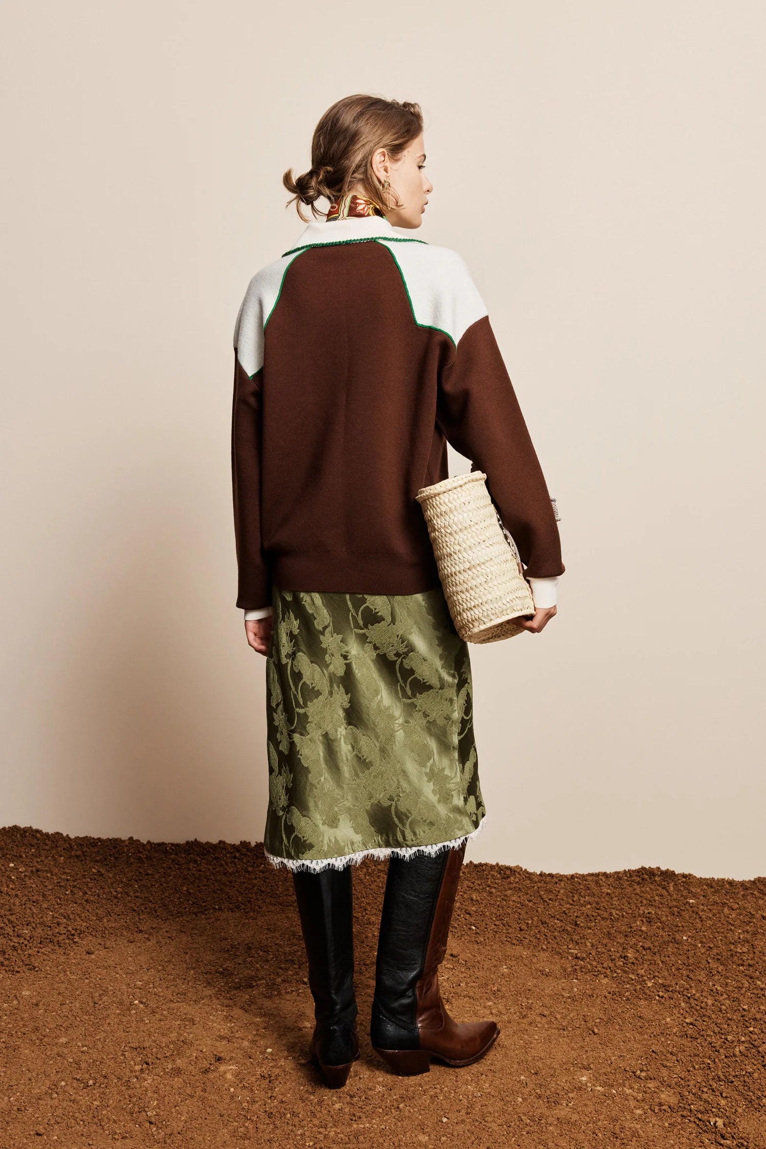 A woman stands with her back to the camera wearing a colorblock knit sweater in brown, white, and green, paired with an olive green patterned skirt, black boots, and holding a woven straw bag against a beige wall and brown floor.
