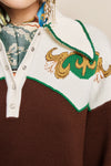 Close-up of a knit sweater with a brown body, white yoke, green trim, and a gold embroidered motif on the chest. A patterned scarf and dangling earrings are partially visible at the neckline.