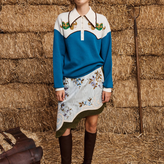 Woman in a blue and white sweater with floral skirt standing in front of hay bales.