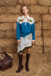 Woman in a blue and white sweater with floral skirt standing in front of hay bales.