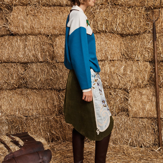 A woman stands in profile against a wall of stacked hay bales, wearing a blue and white color-blocked sweater, an olive green skirt, a white apron with a floral print, and dark brown knee-high boots. A brown leather satchel rests on the straw floor beside her.