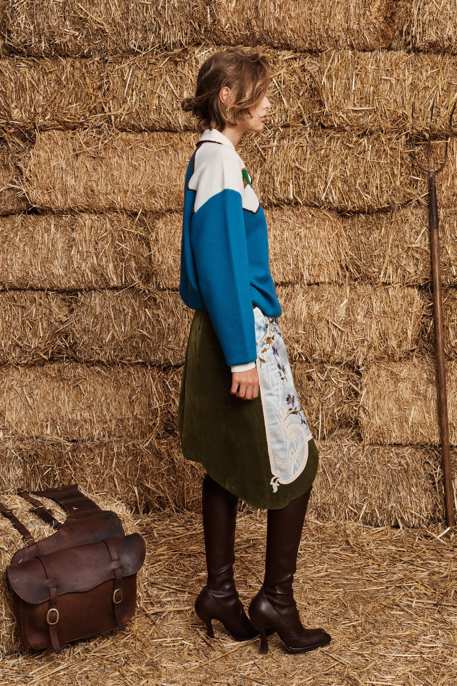 A woman stands in profile against a wall of stacked hay bales, wearing a blue and white color-blocked sweater, an olive green skirt, a white apron with a floral print, and dark brown knee-high boots. A brown leather satchel rests on the straw floor beside her.
