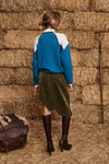 A woman stands with her back to the camera wearing a color-blocked knit sweater with a teal blue body and white shoulder panels, paired with an olive green skirt and dark brown knee-high boots, set against a backdrop of stacked hay bales.