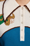 Close-up of a knit polo-style top with a white collar and button placket, blue lower body, brown trim, and a multicolor embroidered motif on the left side.