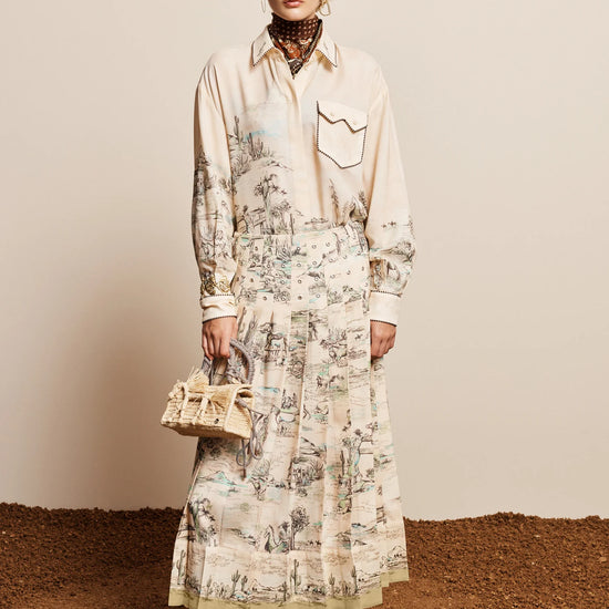 A model stands against a beige wall and brown textured ground, wearing a long-sleeve cream shirt dress with a detailed botanical and landscape print, paired with black ankle boots and holding a small woven handbag.