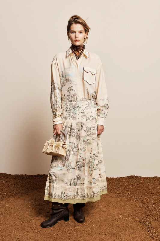 A model stands against a beige wall and brown textured ground, wearing a long-sleeve cream shirt dress with a detailed botanical and landscape print, paired with black ankle boots and holding a small woven handbag.