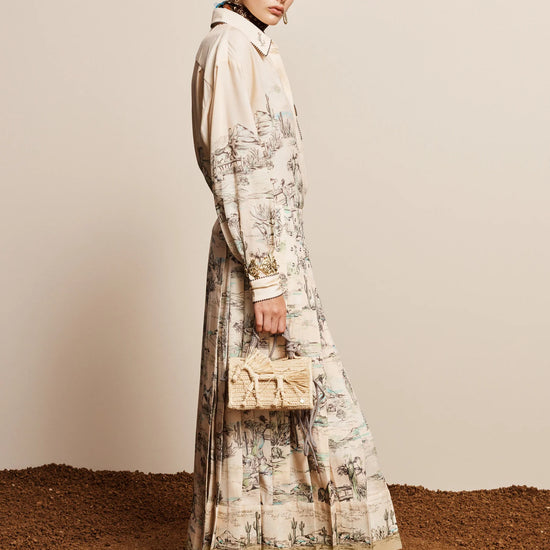 A model stands in profile wearing a long cream-colored shirt dress with a detailed botanical and landscape print, paired with black ankle boots and holding a small beige handbag against a neutral beige wall and textured brown ground.