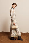 A model stands in profile wearing a long cream-colored shirt dress with a detailed botanical and landscape print, paired with black ankle boots and holding a small beige handbag against a neutral beige wall and textured brown ground.