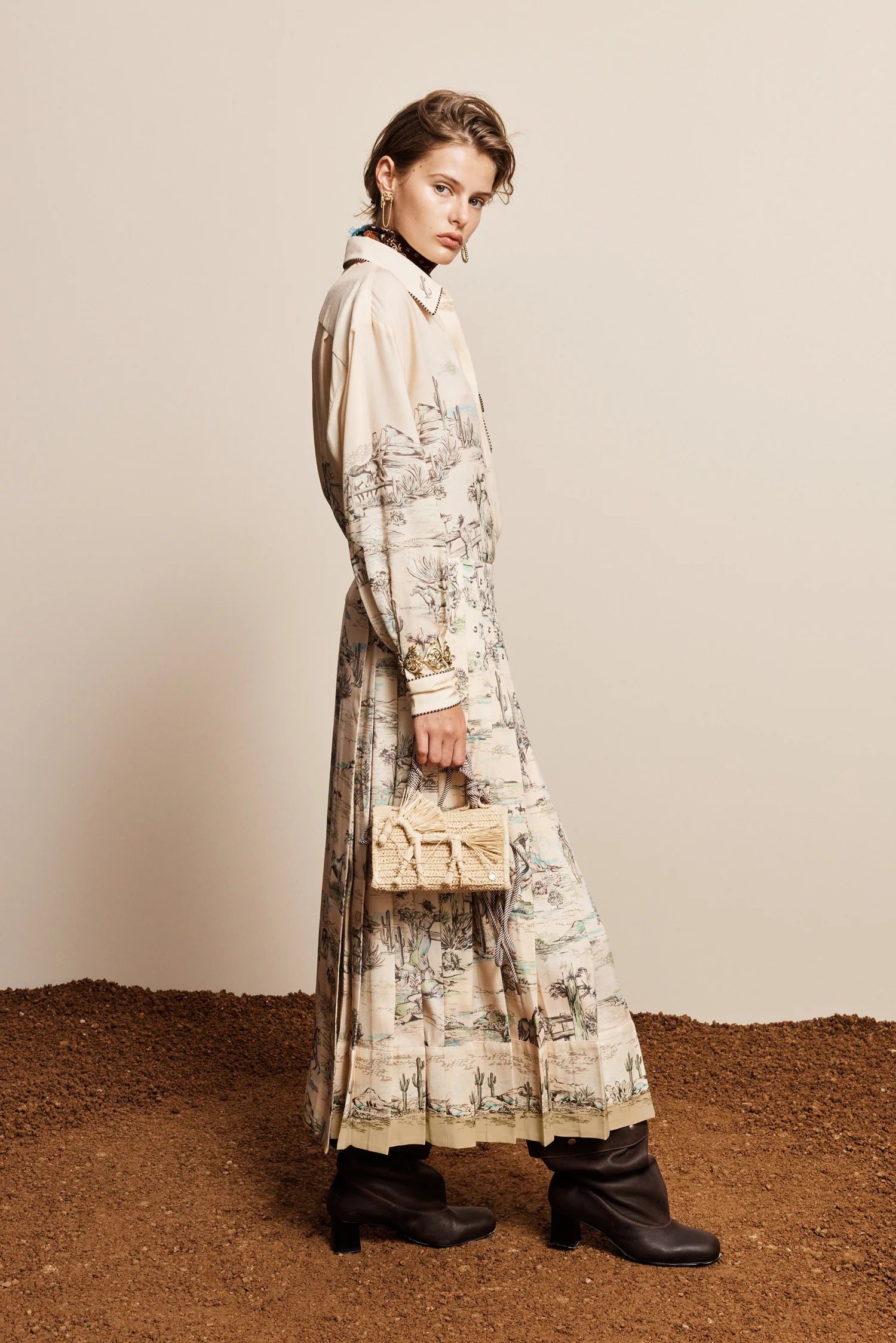 A model stands in profile wearing a long cream-colored shirt dress with a detailed botanical and landscape print, paired with black ankle boots and holding a small beige handbag against a neutral beige wall and textured brown ground.