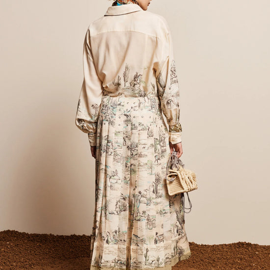 A woman stands with her back to the camera wearing a long cream-colored shirt dress with a beige toile print skirt, holding a small beige handbag against a neutral studio backdrop.