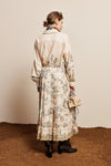 A woman stands with her back to the camera wearing a long cream-colored shirt dress with a beige toile print skirt, holding a small beige handbag against a neutral studio backdrop.