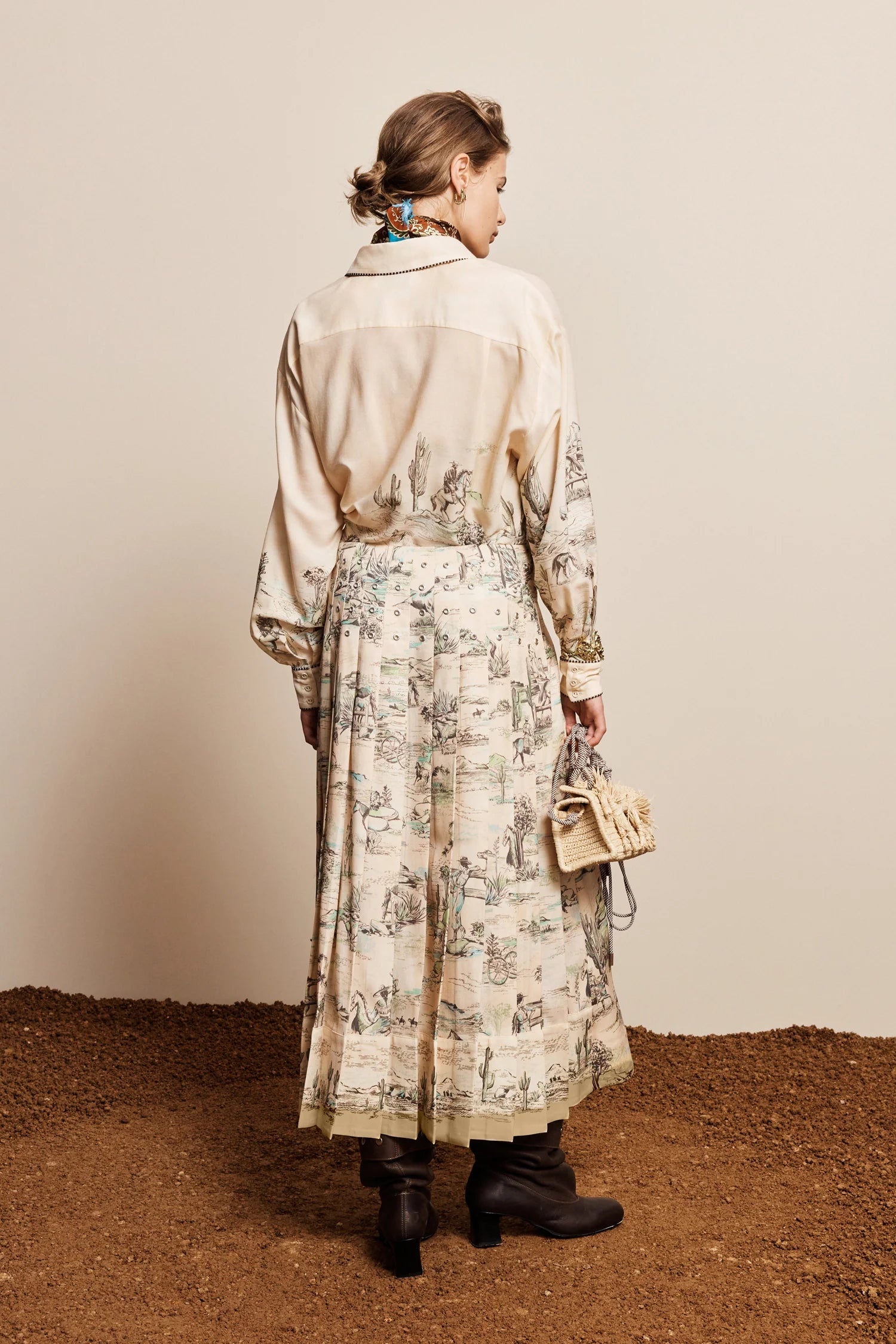 A woman stands with her back to the camera wearing a long cream-colored shirt dress with a beige toile print skirt, holding a small beige handbag against a neutral studio backdrop.