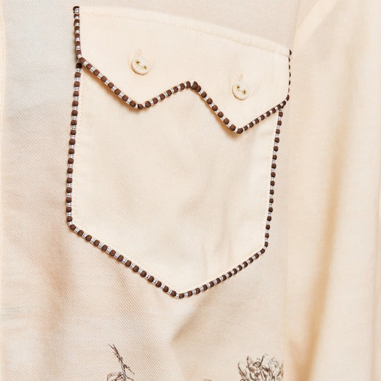 Close-up of a cream-colored shirt featuring a decorative chest pocket with a zigzag flap and dark brown dotted stitching, along with a subtle botanical print near the bottom.