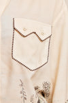 Close-up of a cream-colored shirt featuring a decorative chest pocket with a zigzag flap and dark brown dotted stitching, along with a subtle botanical print near the bottom.