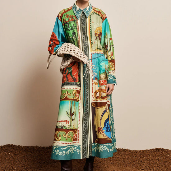 A model stands against a beige studio backdrop wearing a long-sleeve collared coat with a colorful Southwestern desert print, holding a woven straw hat, and wearing black leather boots.