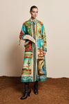 A model stands against a beige studio backdrop wearing a long-sleeve collared coat with a colorful Southwestern desert print, holding a woven straw hat, and wearing black leather boots.