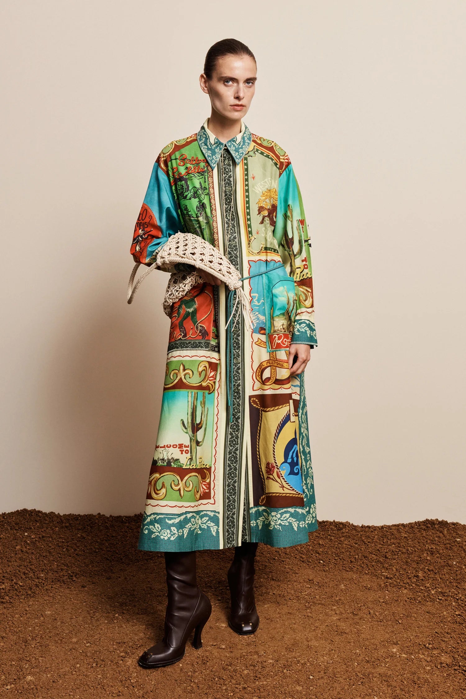 A model stands against a beige studio backdrop wearing a long-sleeve collared coat with a colorful Southwestern desert print, holding a woven straw hat, and wearing black leather boots.