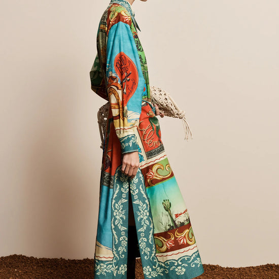 A model is shown from the side in a studio setting, wearing a long-sleeved maxi shirt-dress with a vibrant, multicolored scenic print. The dress features turquoise, green, red, and beige landscape motifs.