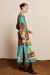 A model is shown from the side in a studio setting, wearing a long-sleeved maxi shirt-dress with a vibrant, multicolored scenic print. The dress features turquoise, green, red, and beige landscape motifs.