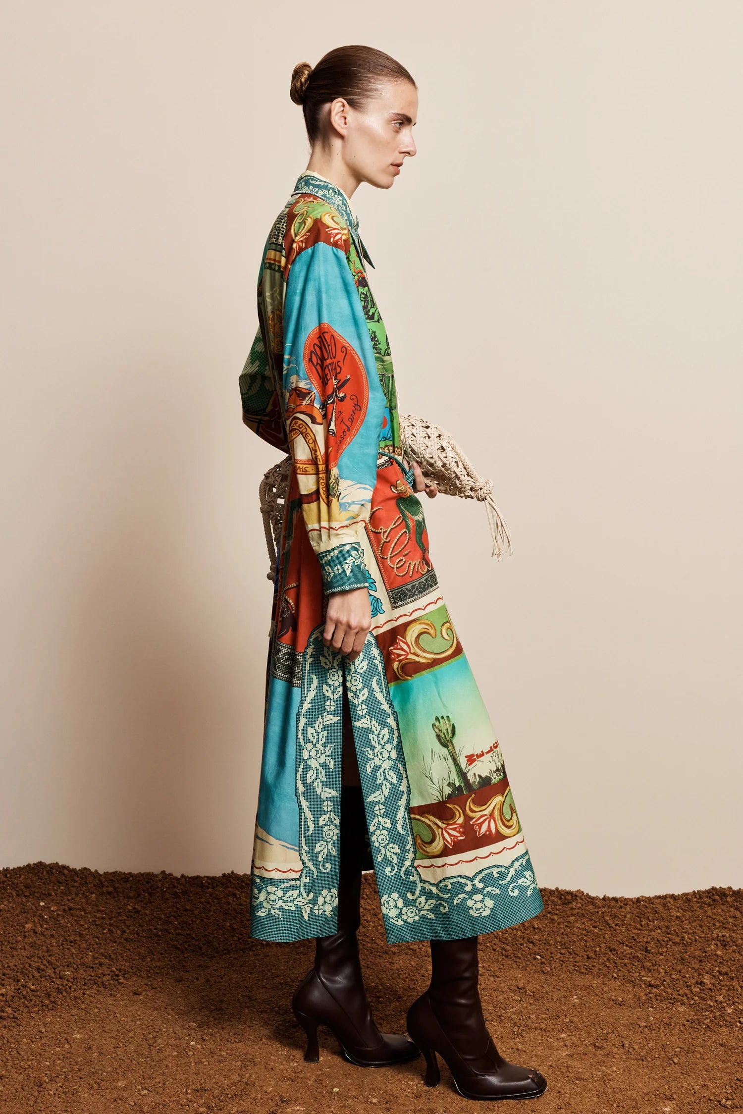 A model is shown from the side in a studio setting, wearing a long-sleeved maxi shirt-dress with a vibrant, multicolored scenic print. The dress features turquoise, green, red, and beige landscape motifs.