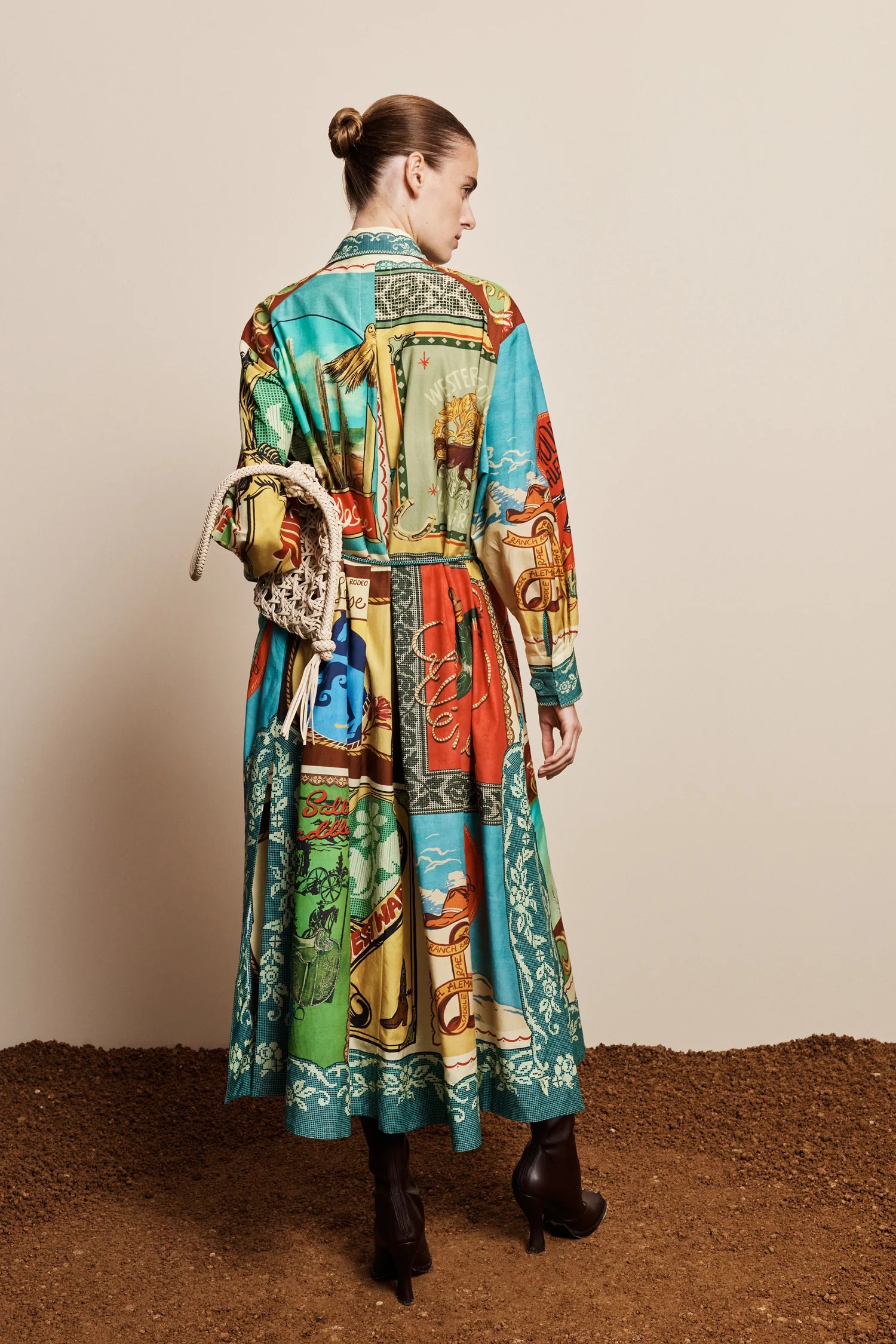 A model is shown from behind wearing a long-sleeved, belted midi dress with a vibrant multicolor patchwork print. The dress features a mix of turquoise, green, red, and yellow panels with a western-themed design.