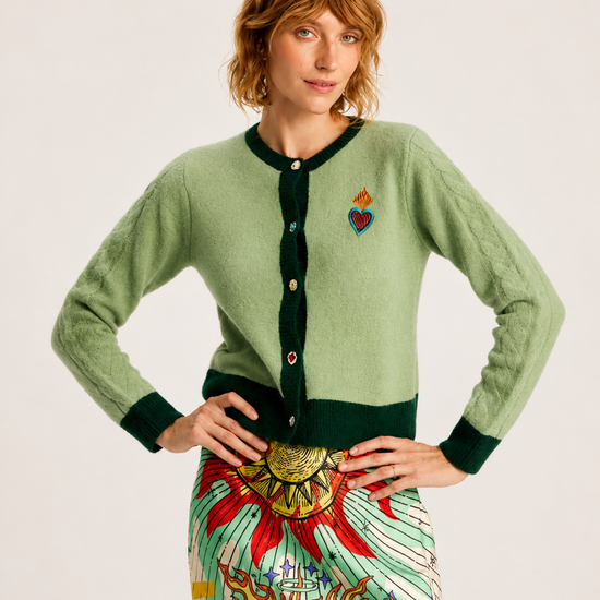 A model wearing a sage green cardigan with dark green trim and a colorful embroidered heart on the chest, paired with a patterned skirt, against a plain light background.