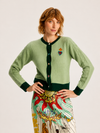A model wearing a sage green cardigan with dark green trim and a colorful embroidered heart on the chest, paired with a patterned skirt, against a plain light background.