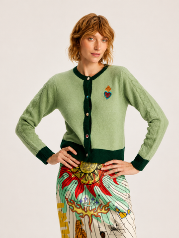 A model wearing a sage green cardigan with dark green trim and a colorful embroidered heart on the chest, paired with a patterned skirt, against a plain light background.