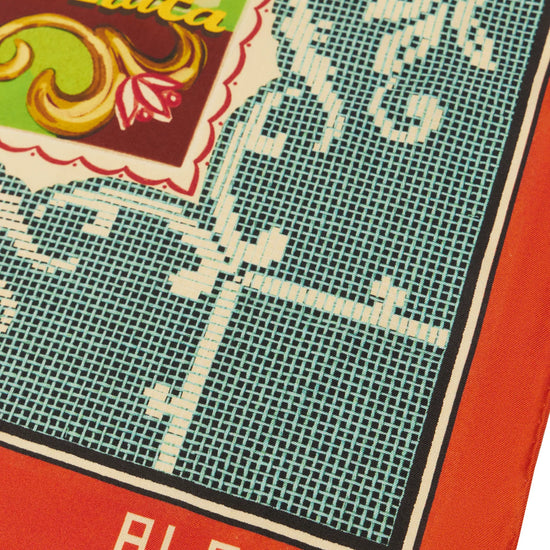 A close-up of a square scarf or bandana featuring a vintage-style print. The design includes a teal grid pattern with white floral scrollwork, a red border, and a decorative multicolor label area with the text 'DEO TO La Plata' and 'ALEMAIS' in white on the red border.