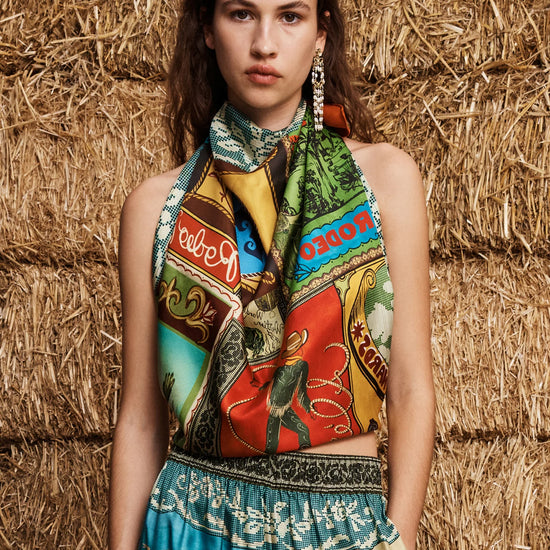 A model wearing a colorful, patterned halter top and matching printed skirt stands in front of a rustic background of stacked straw bales. The garment features a vibrant, multi-colored graphic print with various illustrated motifs and text.