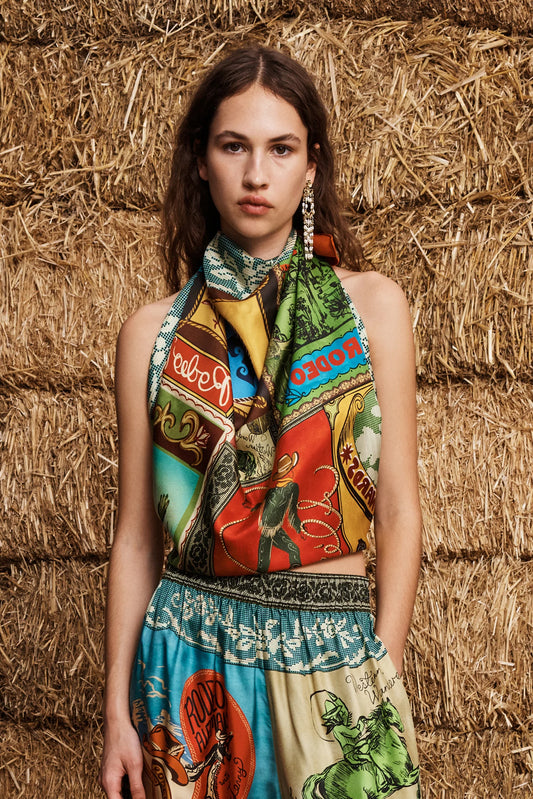 A model wearing a colorful, patterned halter top and matching printed skirt stands in front of a rustic background of stacked straw bales. The garment features a vibrant, multi-colored graphic print with various illustrated motifs and text.