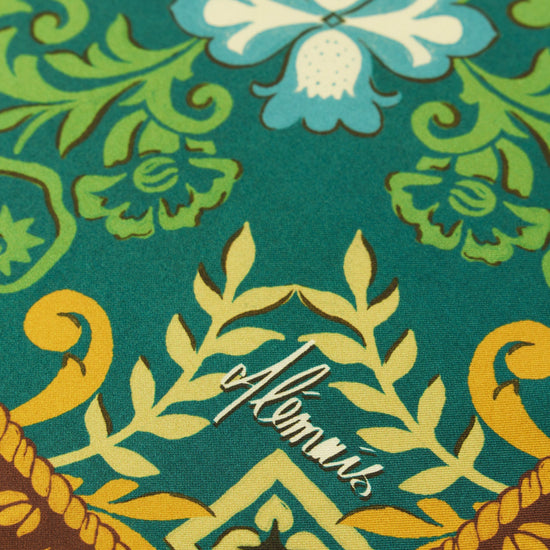 A close-up of a patterned scarf featuring a teal background with green, yellow, and light blue floral motifs, plus a brown decorative border at the bottom.