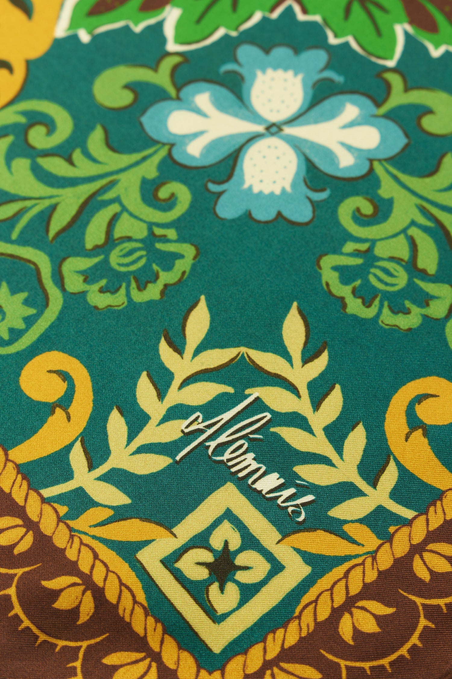 A close-up of a patterned scarf featuring a teal background with green, yellow, and light blue floral motifs, plus a brown decorative border at the bottom.