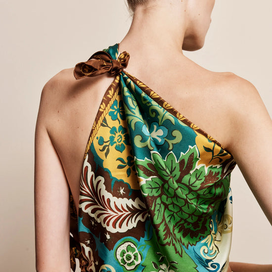 A woman shown from the back wearing a one-shoulder halter top with a vibrant green, yellow, and brown floral print, paired with a cream skirt against a solid beige background.