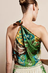 A woman shown from the back wearing a one-shoulder halter top with a vibrant green, yellow, and brown floral print, paired with a cream skirt against a solid beige background.