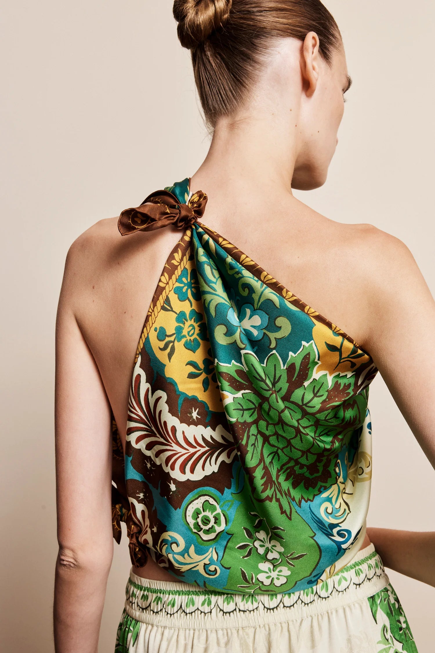 A woman shown from the back wearing a one-shoulder halter top with a vibrant green, yellow, and brown floral print, paired with a cream skirt against a solid beige background.