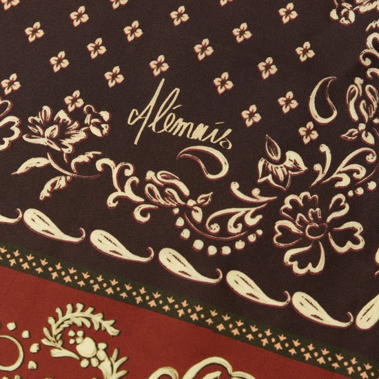 Close-up of a patterned scarf featuring a dark brown upper section with beige floral motifs and the script brand name  and a red lower section with beige ornamental paisley and border details.