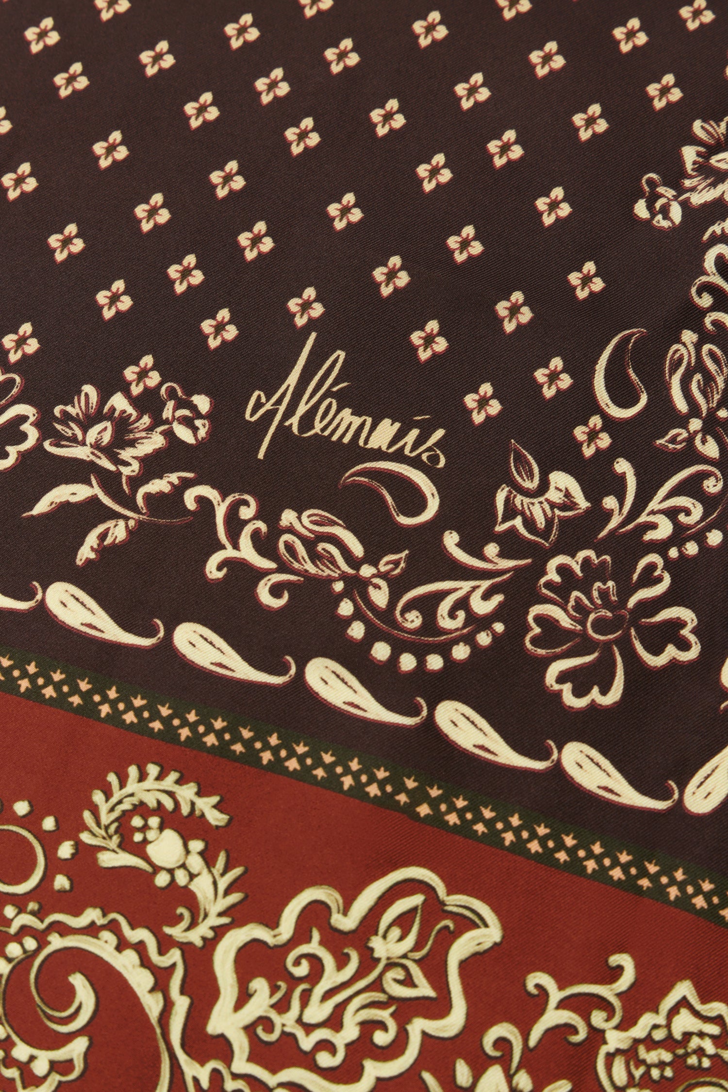 Close-up of a patterned scarf featuring a dark brown upper section with beige floral motifs and the script brand name  and a red lower section with beige ornamental paisley and border details.