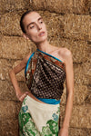 A woman posing in front of stacked hay bales, wearing a two-piece printed outfit consisting of a brown patterned draped top and a cream-colored skirt with green floral prints.
