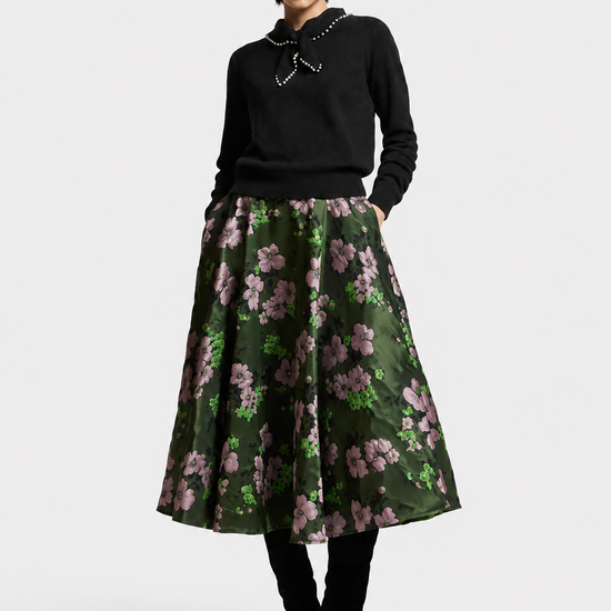A model stands against a light gray studio background wearing a black knit sweater with a silver collar, a dark green A-line skirt with a pink and green floral print, black tights, and black heeled boots.