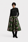 A model stands against a light gray studio background wearing a black knit sweater with a silver collar, a dark green A-line skirt with a pink and green floral print, black tights, and black heeled boots.