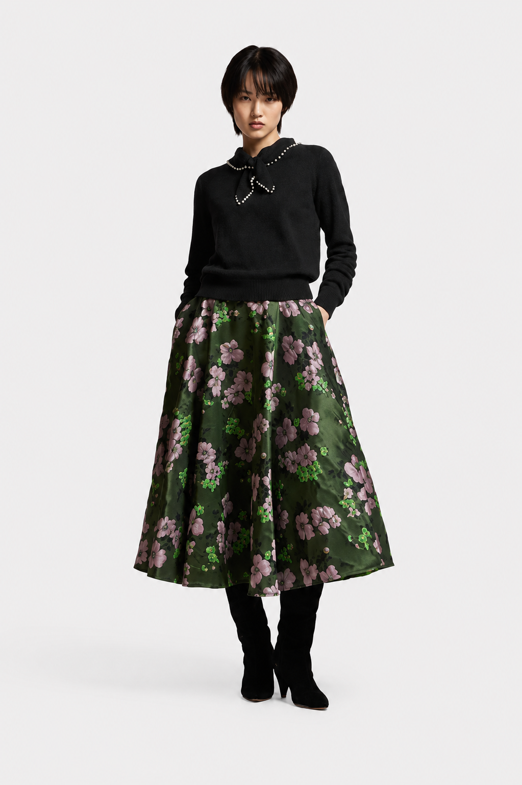 A model stands against a light gray studio background wearing a black knit sweater with a silver collar, a dark green A-line skirt with a pink and green floral print, black tights, and black heeled boots.