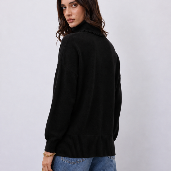 A woman wearing a black knit sweater with a high ruffled mock neck, shown from a three-quarter back view against a light gray studio background.